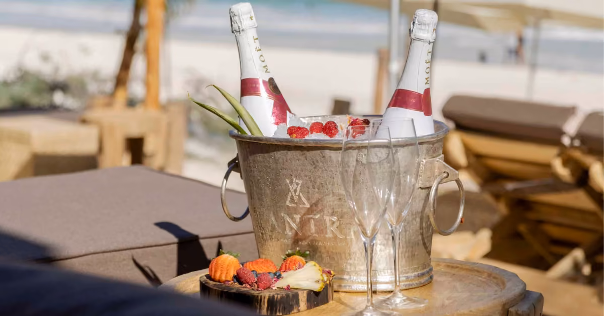 A silver ice bucket with two Moët Chandon bottles and raspberries sits on a table at the beach, flutes and fruit nearby.