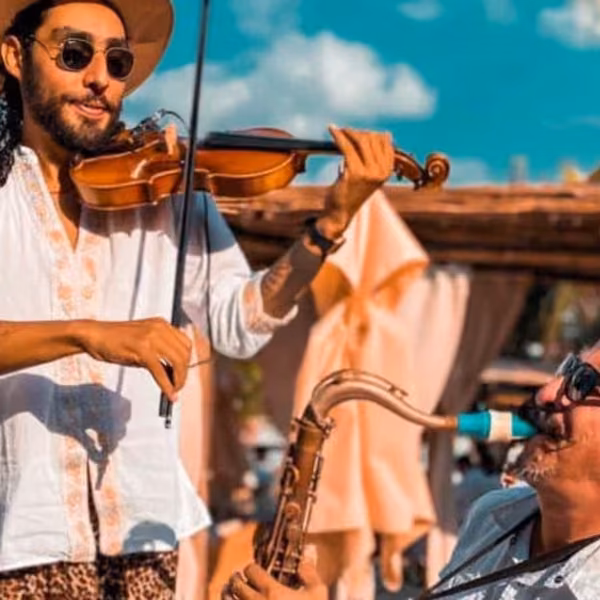 Two musicians play violin and saxophone outdoors at Tantra Beach Club, wearing sunglasses, with palm trees and sky behind them.