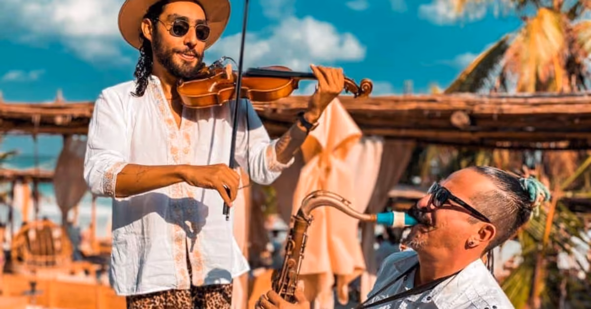 Two musicians play violin and saxophone outdoors at Tantra Beach Club, wearing sunglasses, with palm trees and sky behind them.