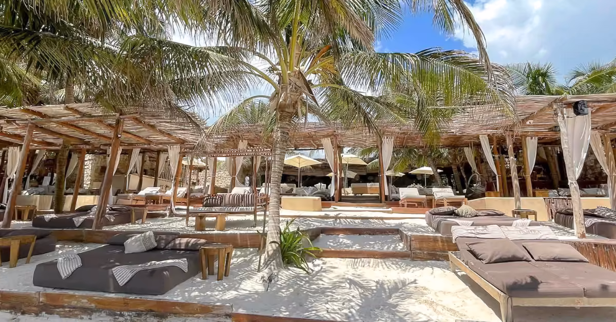 Beach lounge at Tantra Beach Club with shaded cabanas, daybeds, small tables, and palm trees on white sand under a sunny sky.