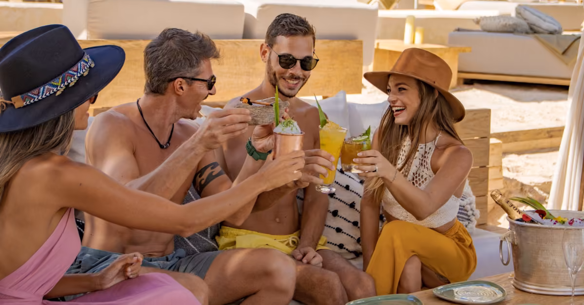 Four friends in summer outfits toast with colorful drinks at a beach club, smiling by lounge chairs and a fruit basket.