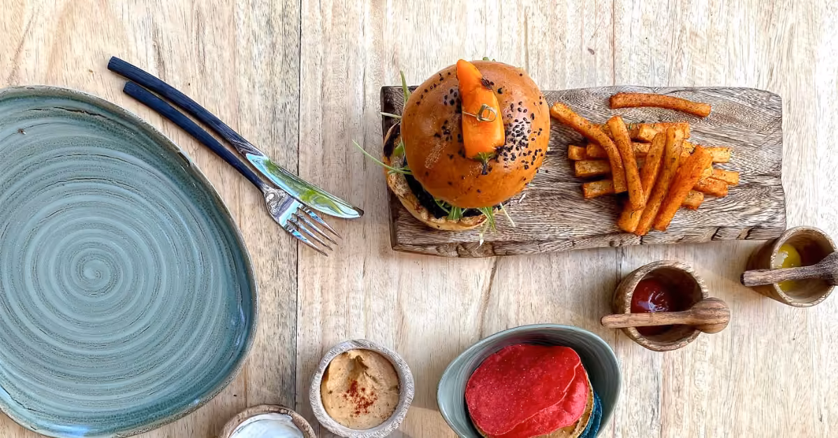 Burger and fries on a wooden board next to an empty green plate, fork, knife, dips, ketchup, mustard, and chips.