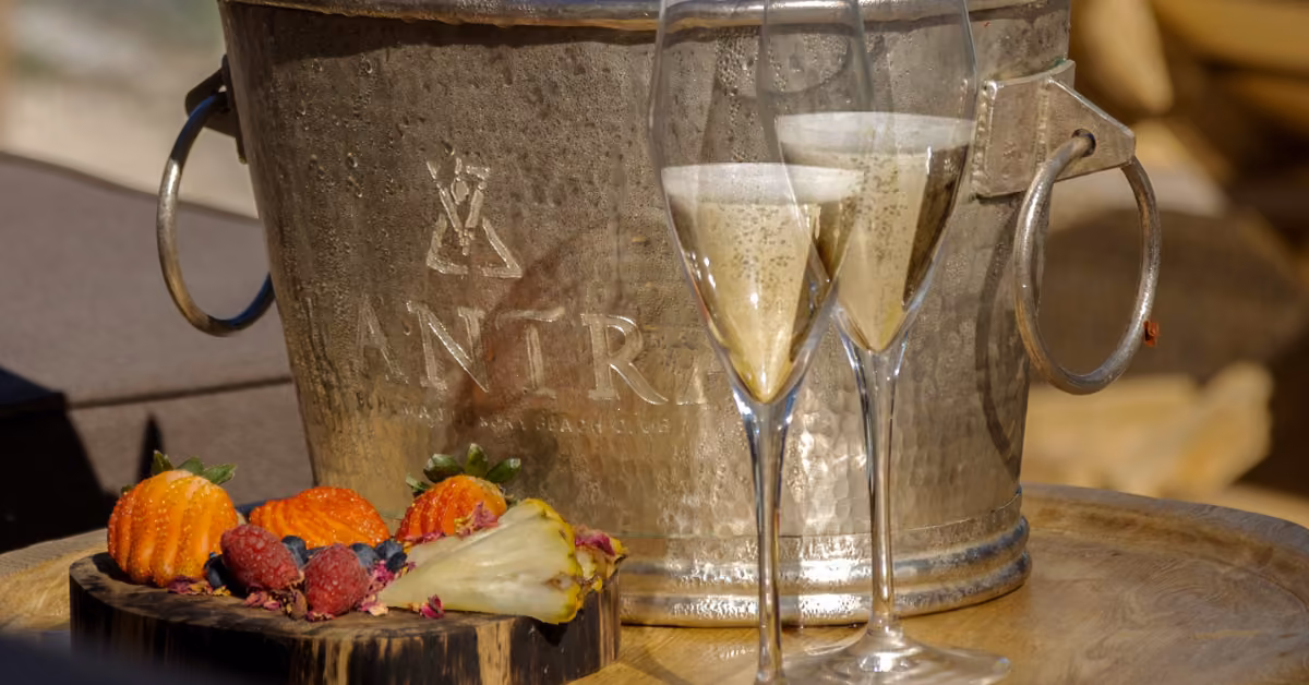 Two glasses of sparkling wine on a wooden tray with champagne and a plate of mixed fruits, including strawberries and pineapple.