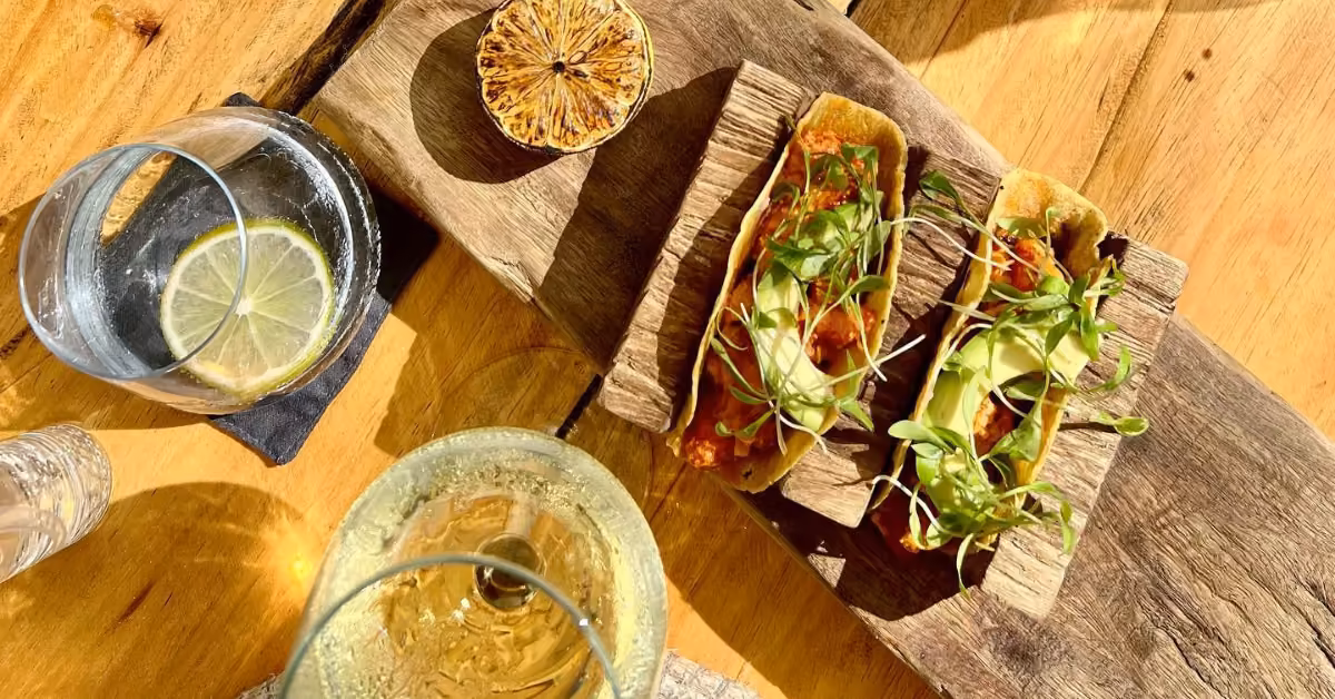 Two tacos with greens and avocado on a wooden board, served with grilled lemon, a lemon drink, and white wine at a beach club.