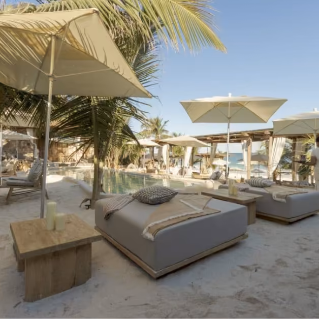 Beach lounge at Tantra with cushioned beds, wooden tables, umbrellas, palm trees, pool, and ocean views under a blue sky.