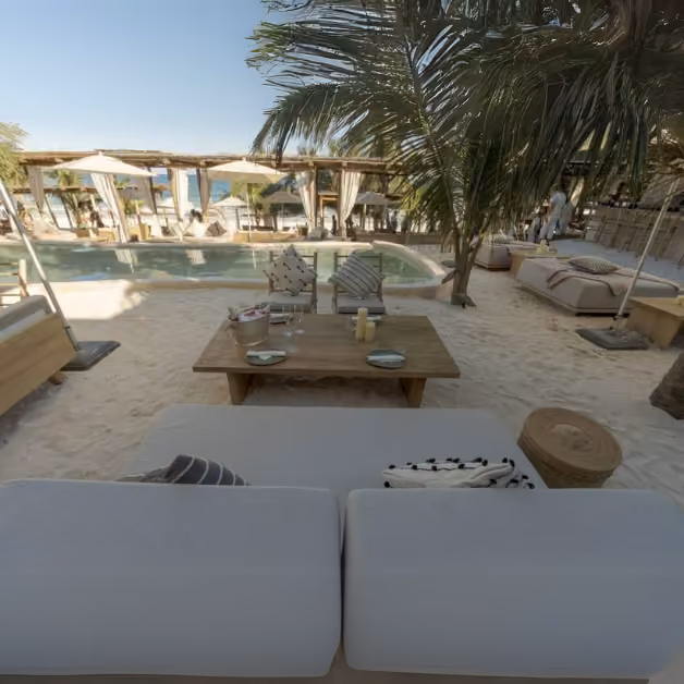 White cushioned lounge seating and a wooden table on sandy ground by a pool, with palm trees and cabanas under blue sky.