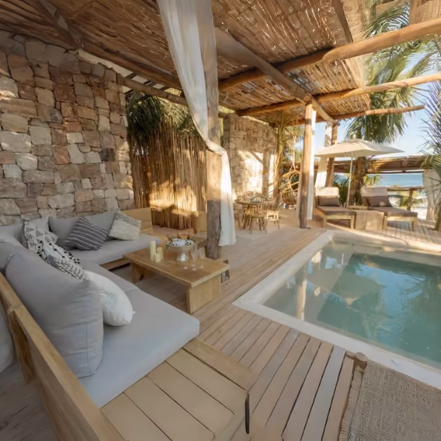 Tantra Beach Club’s outdoor lounge has wood furniture, beige cushions, a small pool, bamboo roof, palms, and ocean views.