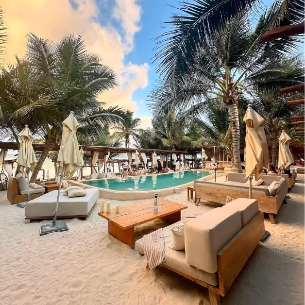 Chic beachside lounge with cushioned sofas, sunbeds, umbrellas on white sand by a pool, framed by palm trees and clouds.