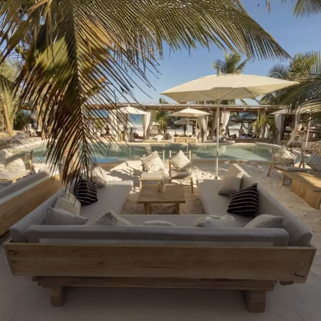An inviting lounge with cushioned sofas overlooks a pool, palm trees, and umbrellas, with the beach and ocean in the background.