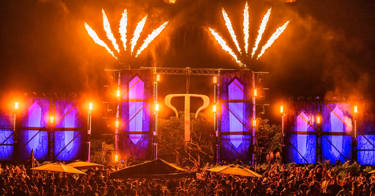 A lively crowd enjoys the tehmplo music festival at night, watching a bright blue-lit stage with fireworks in the sky.