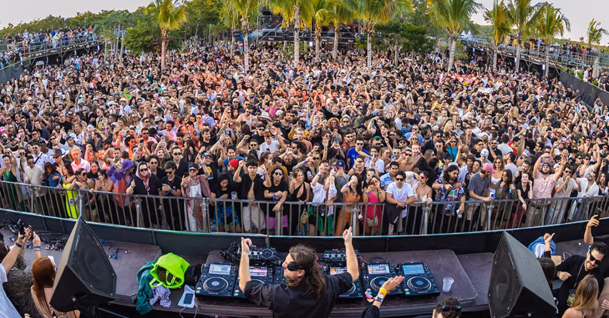 A DJ plays on stage as a lively crowd dances and cheers beneath palm trees at an outdoor festival under a sunny sky.