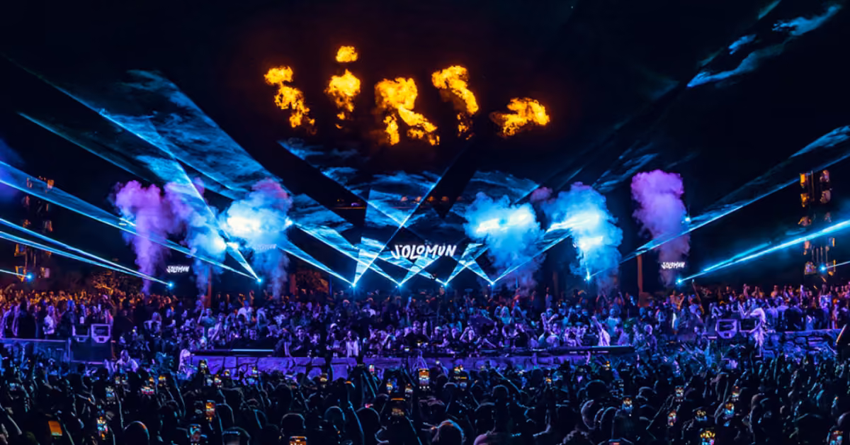 A crowd at Tehmplo festival faces a stage with blue lasers, smoke, and fire effects above the DJ, creating a lively night scene.