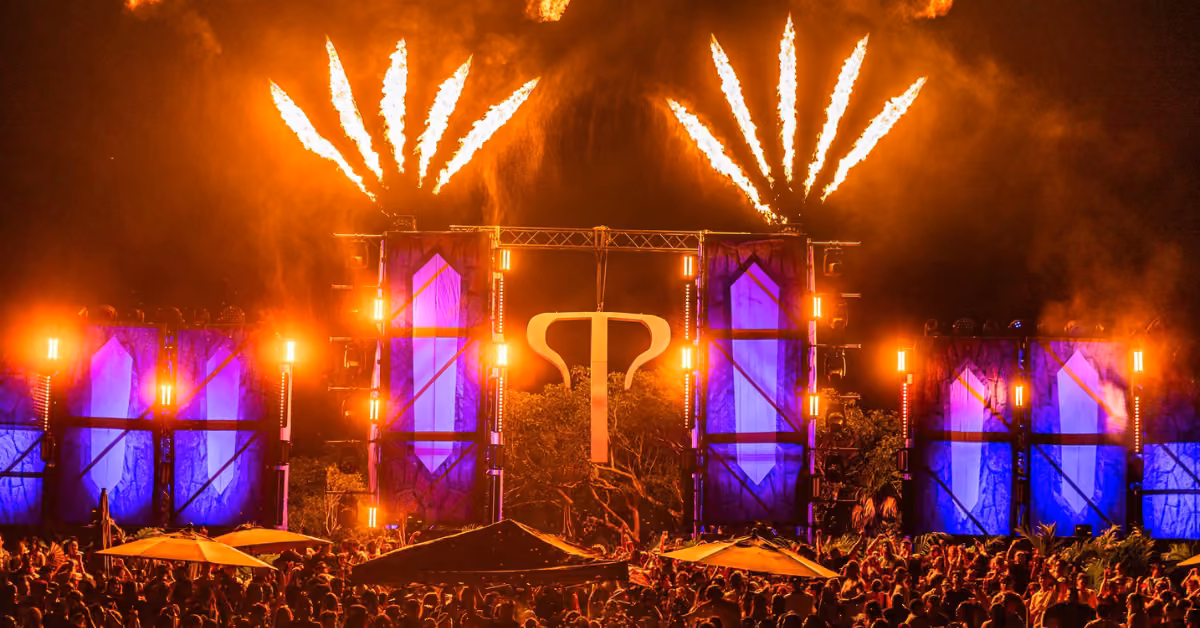 A lively crowd at Tehmplo festival watches orange fireworks and lights illuminate a stage with a glowing symbol and blue panels.