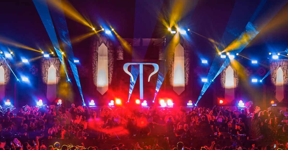 A large crowd enjoys a nighttime music festival with colorful stage lights and a glowing symbol projected behind the performers.
