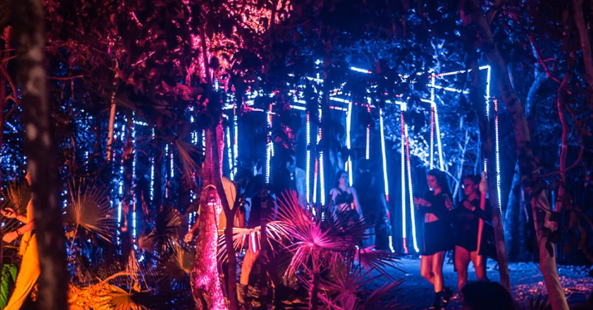Festival-goers stroll through a forest lit by pink, purple, and blue neon lights, creating a lively nighttime atmosphere.