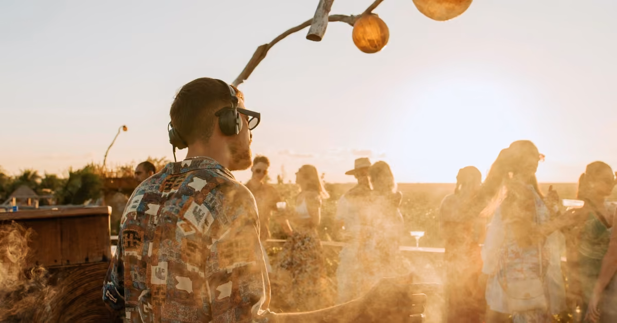 A man in headphones and a patterned shirt stands on a sunlit rooftop as people enjoy a smoky sunset gathering around him.