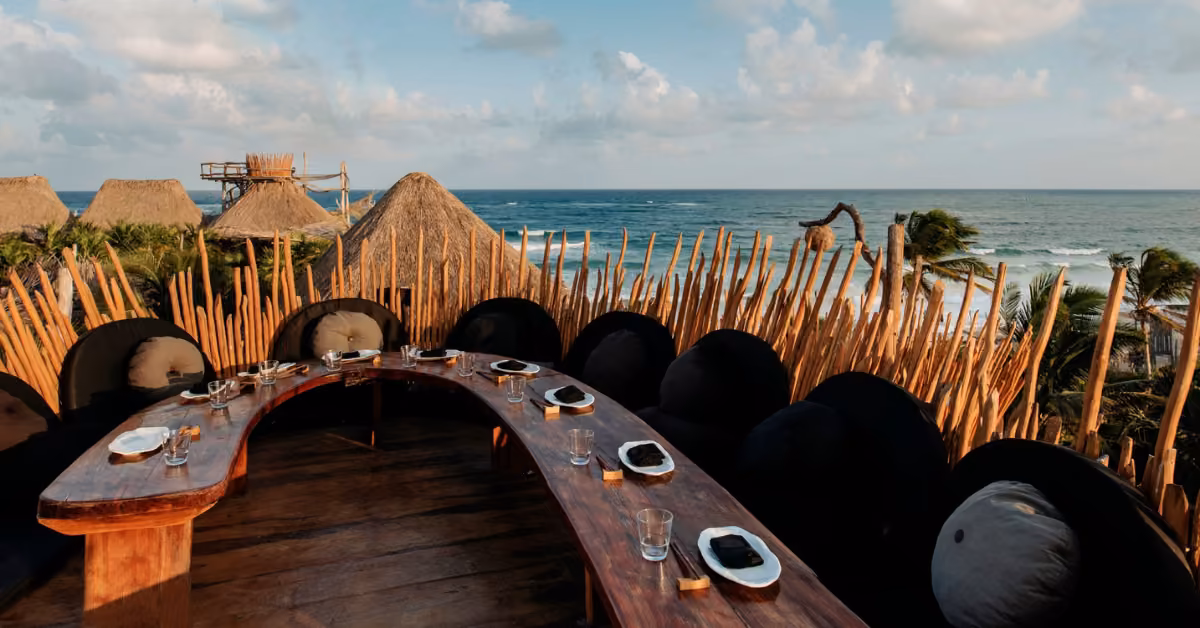 Curved wooden dining table with plates and glasses, cushioned chairs, tropical beach view, thatched huts, palm trees, clouds.