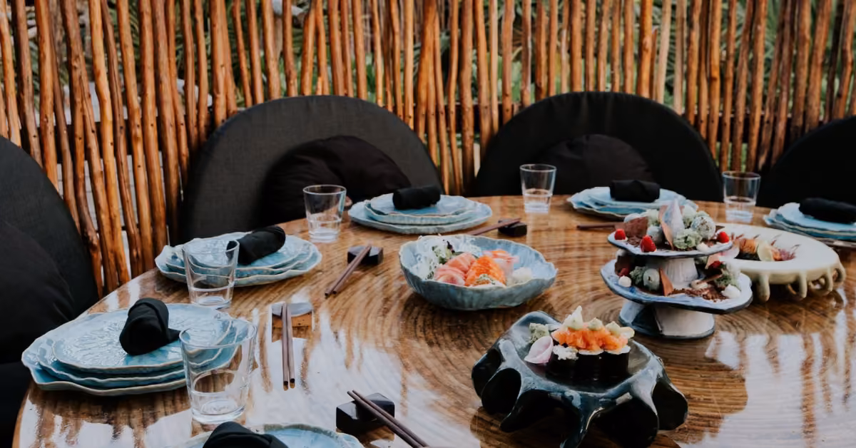 A round wooden table with blue dishes, black napkins, sushi platters, and glasses on a rooftop with chairs and bamboo fence.
