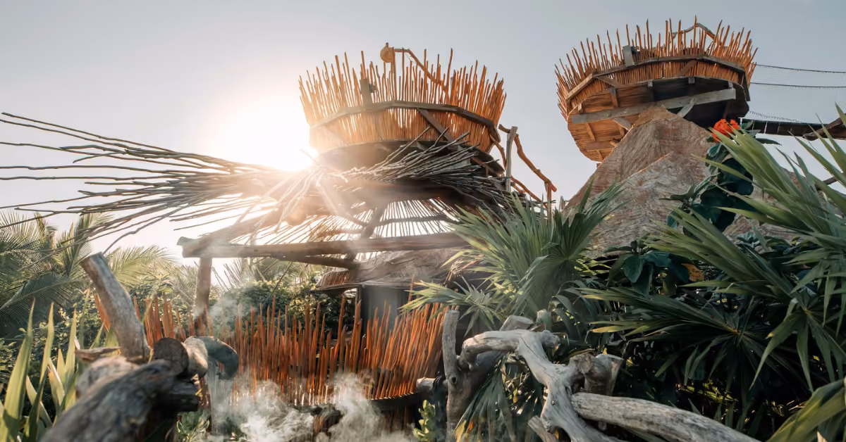 Wooden structures with spiky tops sit among lush plants on a rooftop in sunlight, mist rising under a clear sky.