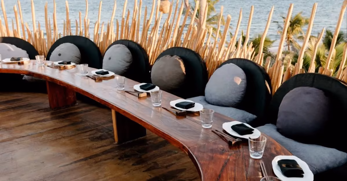 Curved wooden dining table with place settings and large cushions on a rooftop, overlooking water and palm trees.