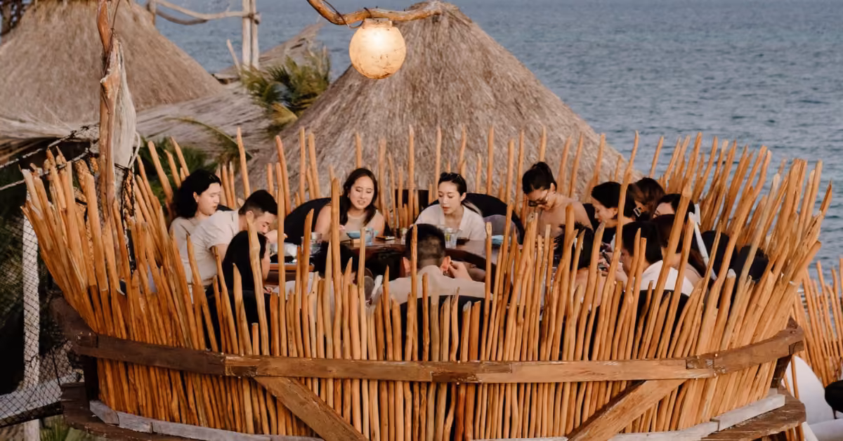 People share a meal at a round wooden nest-like table on a rooftop, with thatched roofs and the ocean visible in the background.