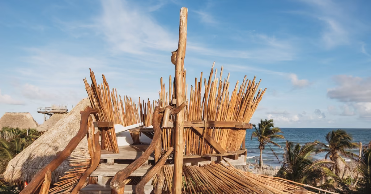 A rustic wooden structure with tall sticks faces palm trees and the ocean, with thatched roofs in the background under a blue sky.