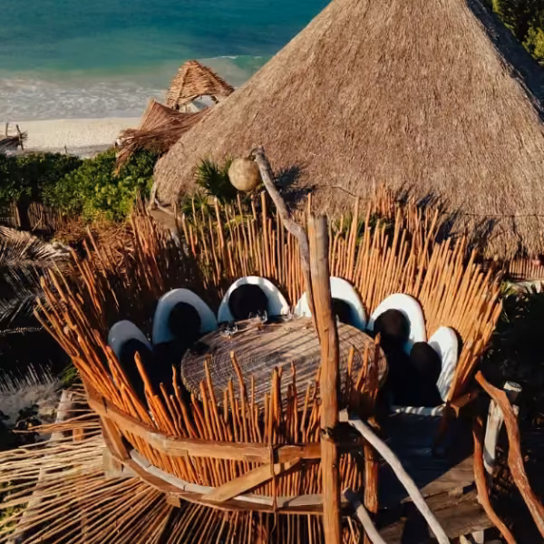 Rustic rooftop dining area with circular table and curved chairs, under a thatched roof, overlooking beach and turquoise sea.