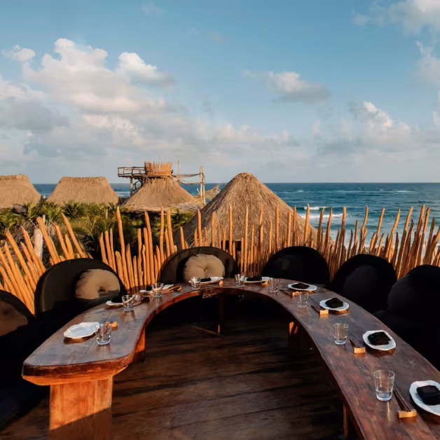 Curved wooden dining table with plates and glasses on a rooftop terrace overlooking the ocean, huts, and a cloudy sky.