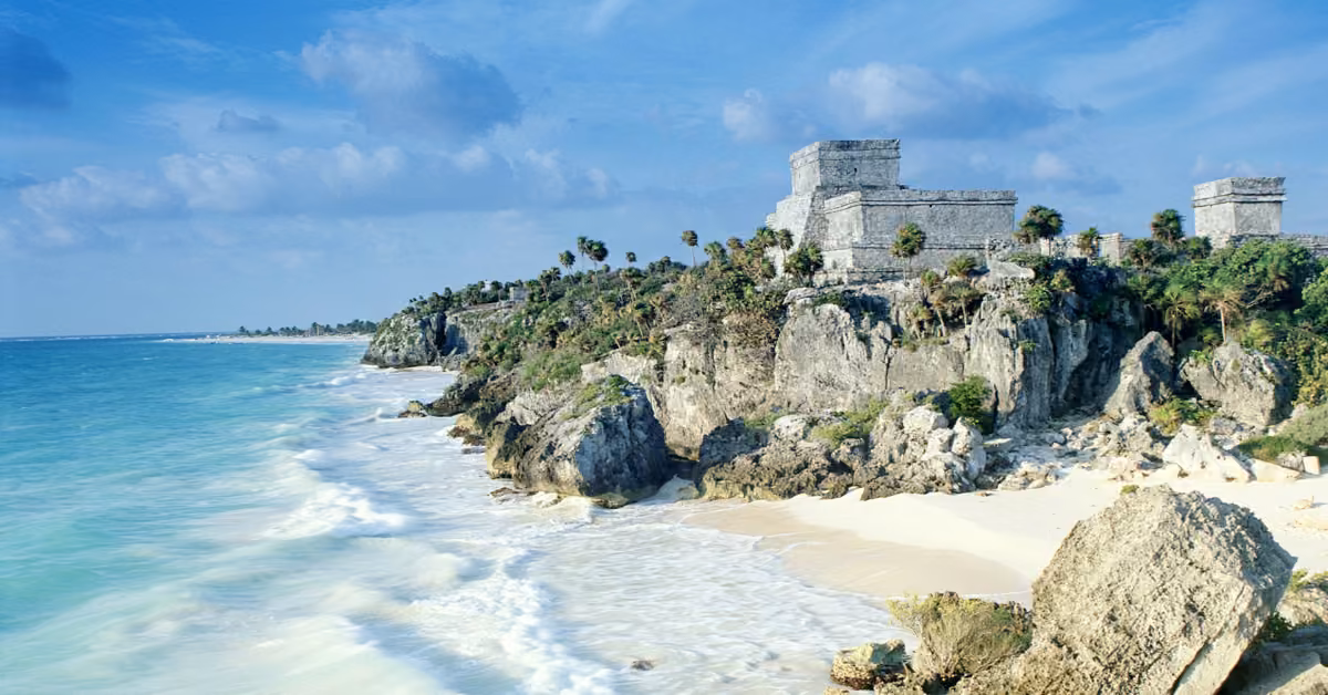Mayan ruins sit on a rocky cliff above turquoise sea and white sand, palm trees dotted around under a partly cloudy sky.