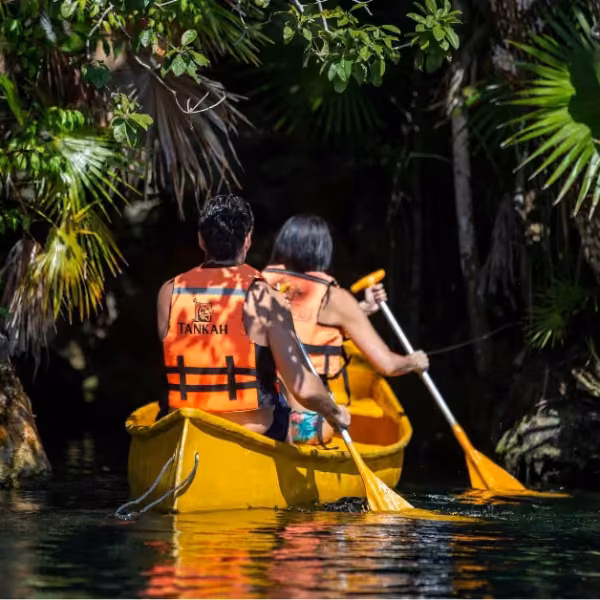 Two people in orange life jackets paddle a yellow canoe through lush green foliage on a calm tropical river.