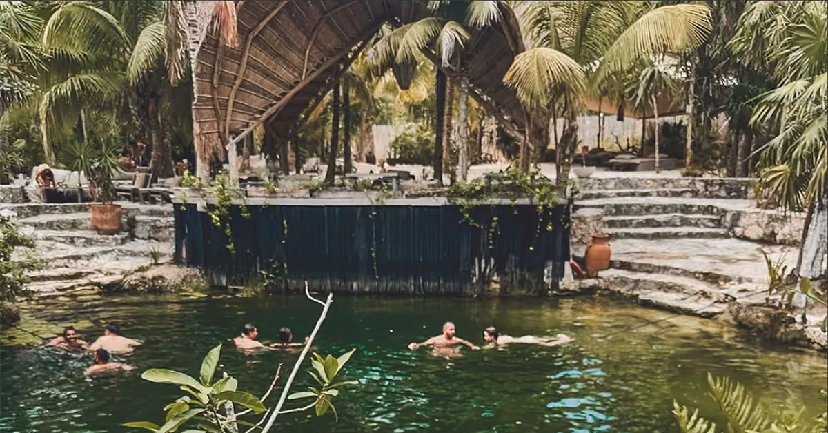 People swim and lounge in Vesica Cenote Club’s green pool, surrounded by stone steps, palm trees, and a thatched shelter.