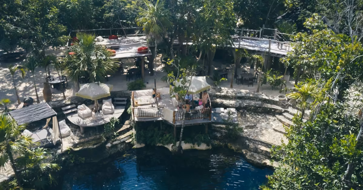 Vesica Cenote Club with tables and umbrellas overlooking a blue cenote, surrounded by lush trees and walkways in nature.