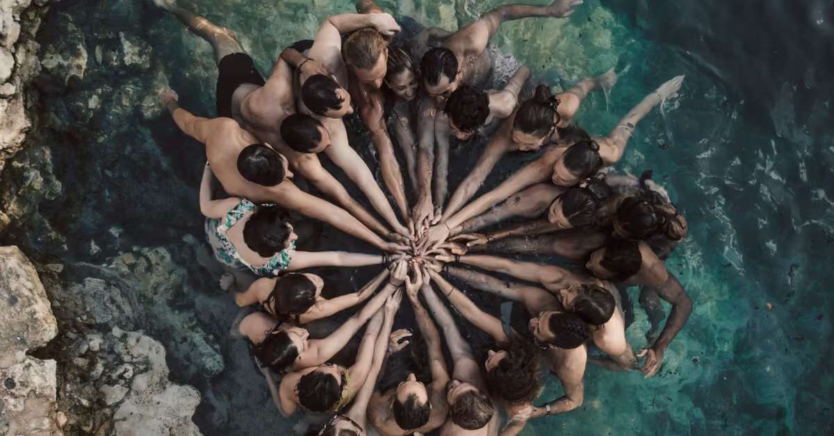 People stand in a clear cenote, forming a circle and reaching to touch hands at the center, creating a radial pattern from above.