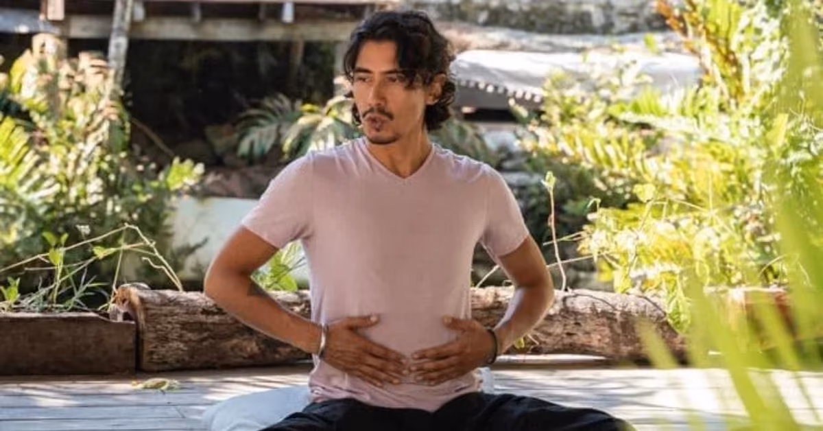 A dark-haired man meditates cross-legged on a wooden platform outdoors, hands on his abdomen, surrounded by lush greenery.