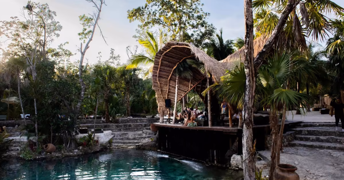 People lounge beneath a curved thatched roof at Vesica Cenote Club beside a pool, stone steps, and lush jungle greenery.