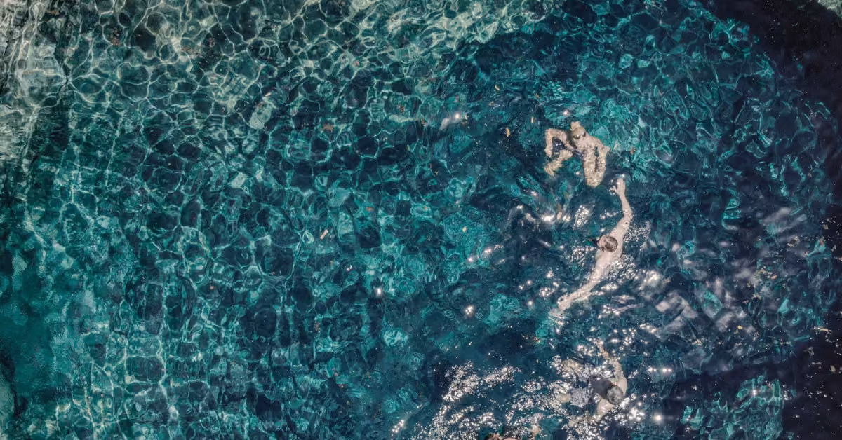 Two people swim in clear blue-green water at Vesica Cenote Club, sunlight dancing on the gentle ripples around them.