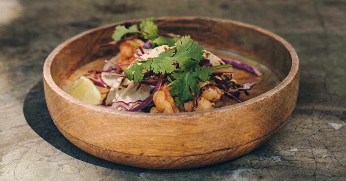A wooden bowl with shrimp, red cabbage, cilantro, and lime wedges sits on a textured gray surface with fresh ingredients.