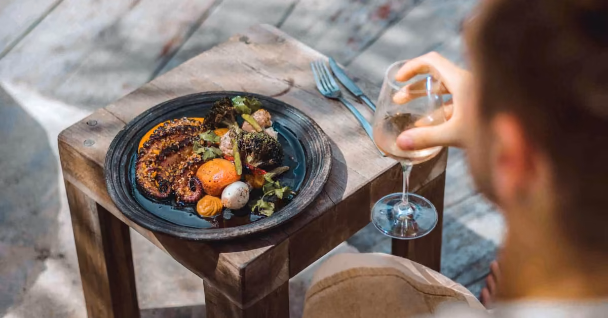 A person at a rustic table enjoys grilled vegetables and octopus, holding a glass of white wine beside their utensils.