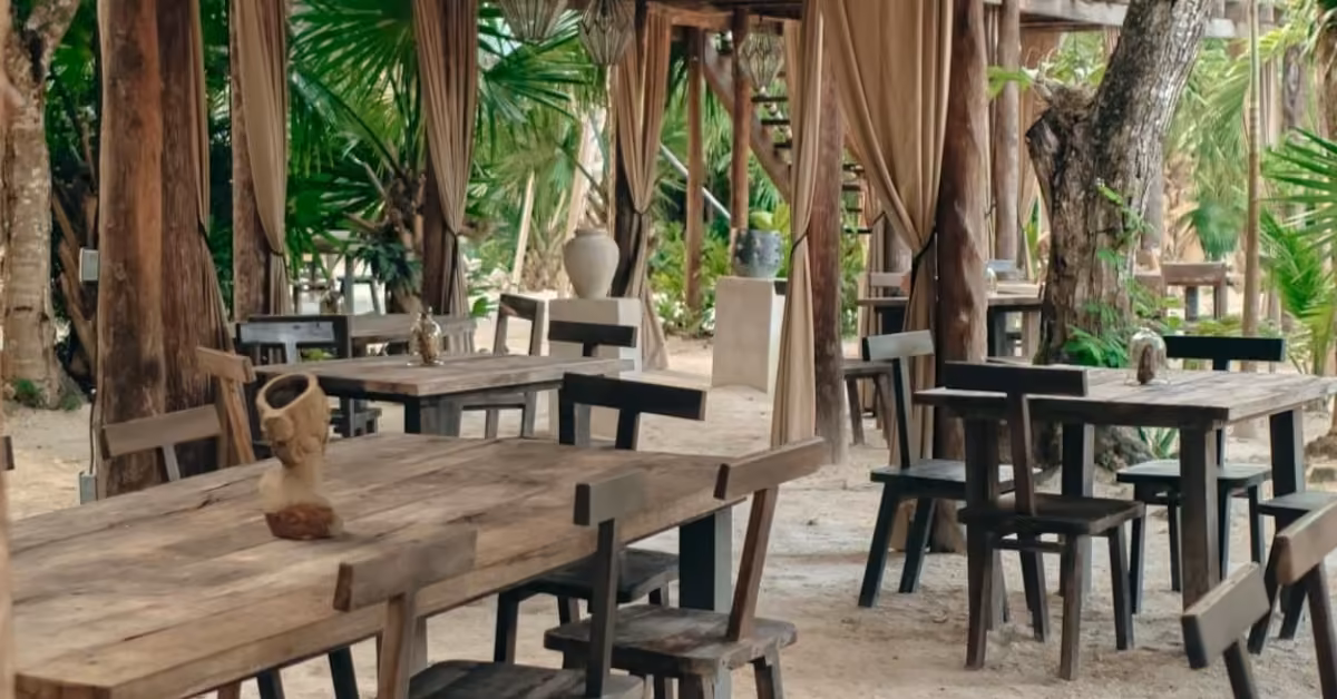 Rustic wooden tables and chairs on sandy ground, framed by palm trees and beige curtains for a relaxed, tropical vibe.