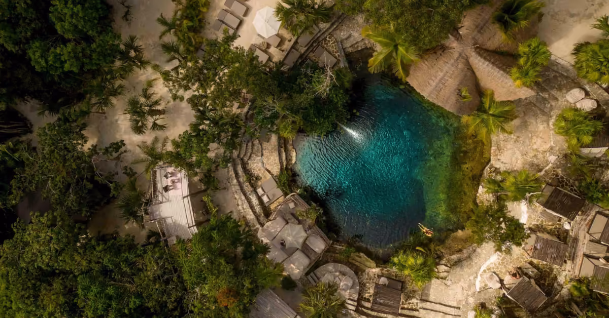 Tropical lagoon at Vesica Cenote Club with clear blue water, lush greenery, wooden huts, sandy paths, loungers, and walkways.