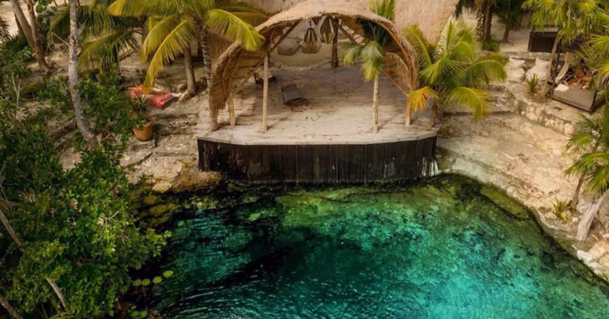 A clear turquoise lagoon borders Vesica Cenote Club’s thatched-roof deck and hammocks, set among rocks and palm trees.