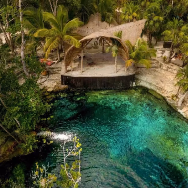 Thatched-roof stage shaded by palms beside a turquoise cenote, surrounded by lush greenery and rocky edges.