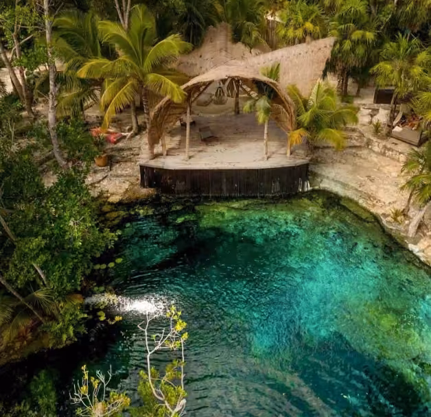 Thatched-roof stage shaded by palms beside a turquoise cenote, surrounded by lush greenery and rocky edges.