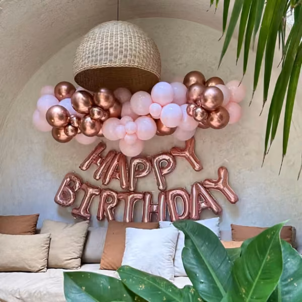 A cozy seating area with beige and brown cushions, pink and rose gold balloons, a wicker lamp, and lush green plants.