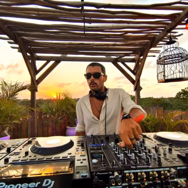 A DJ in sunglasses performs at sunset on an outdoor deck, surrounded by plants and wood decor, mixing music at his setup.
