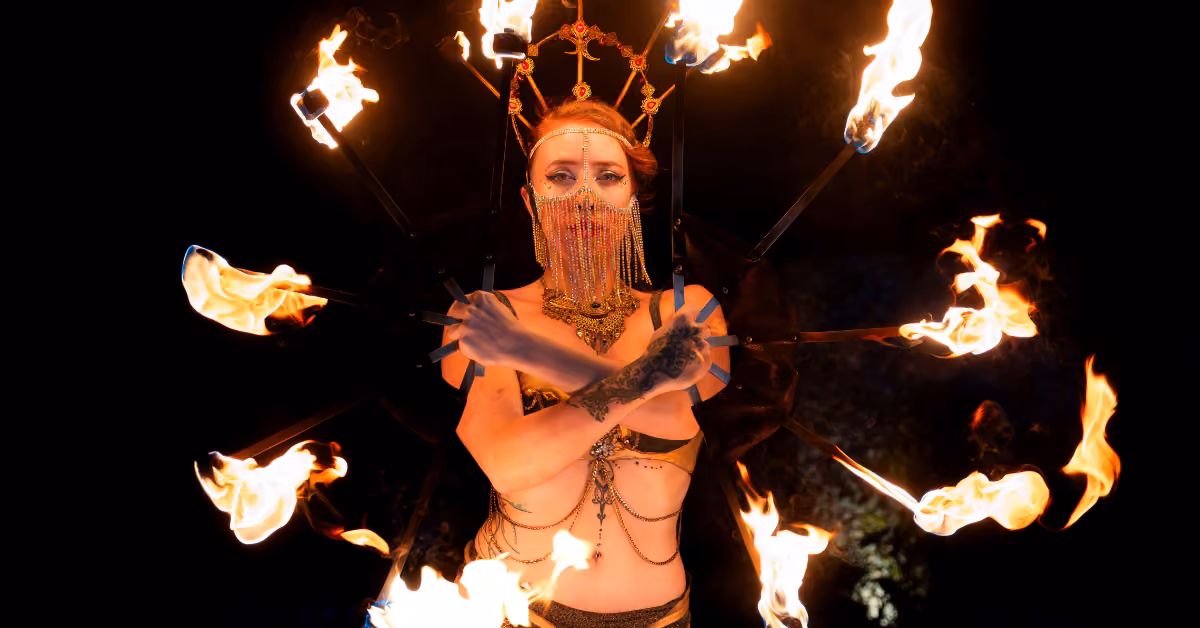 Performer in gold attire and beaded mask stands arms crossed, ringed by flaming torches against a dark background.