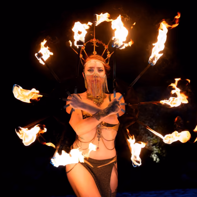 A performer in a gold costume and ornate headpiece holds a flaming circular prop against a dark background.