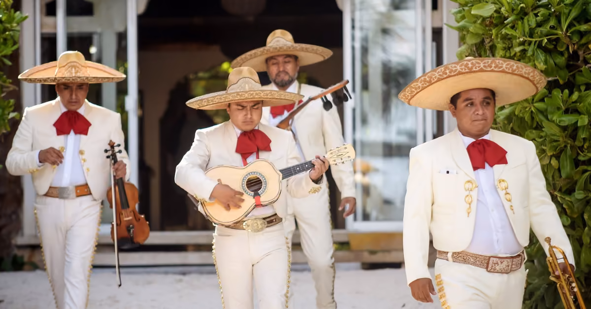 Four mariachi musicians in white suits and sombreros walk outside. One plays guitar, another has a violin. Plants and a doorway behind.