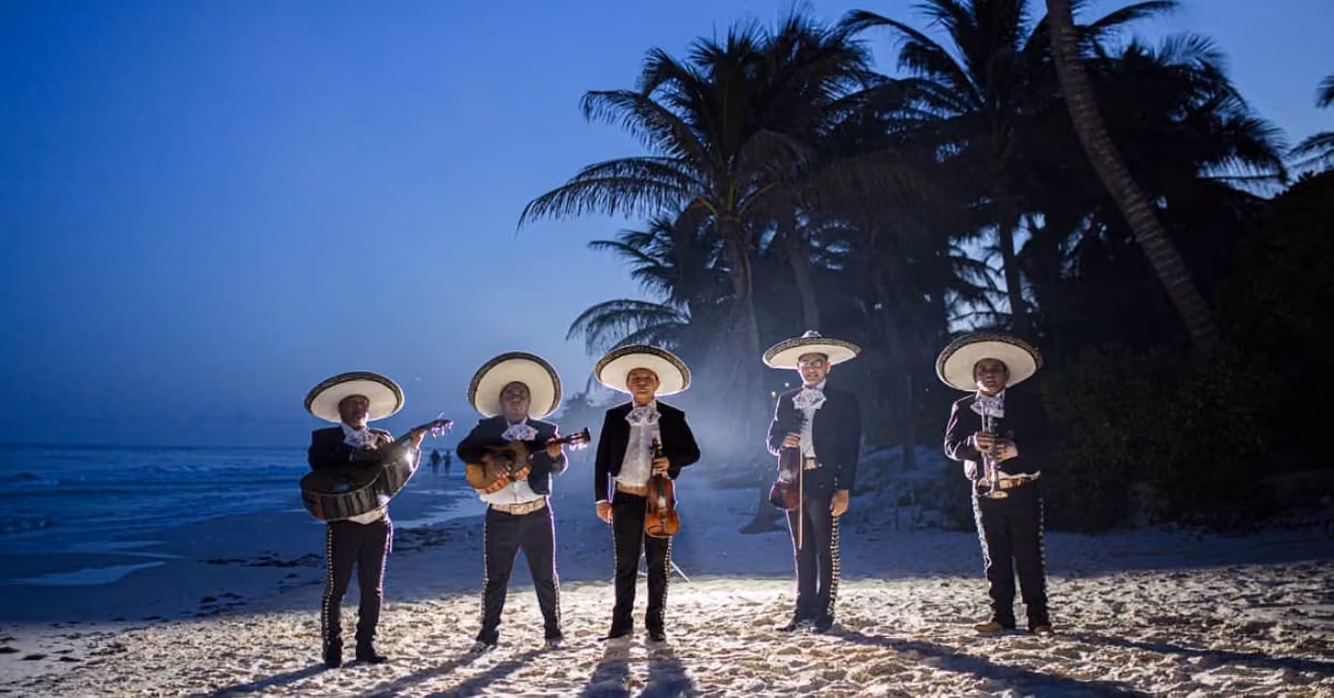 Five mariachi musicians in traditional outfits and sombreros play on a beach at dusk, with palm trees and the ocean behind them.