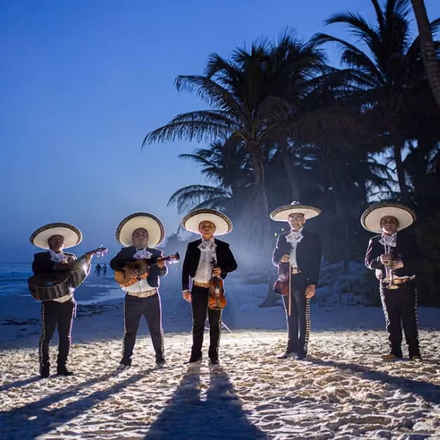 Five mariachi musicians in traditional outfits and sombreros stand on a sandy beach at dusk, ocean and palms behind them.